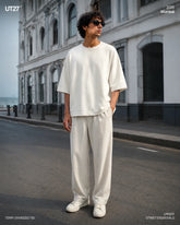 Full-length fashion editorial shot of a male model standing on an urban street in Mumbai, wearing an off-white heavyweight Terry oversized t-shirt and matching relaxed-fit wide-leg trousers. He is wearing dark sunglasses and white sneakers, posing against a classic colonial white building facade. High-end streetwear lookbook photography by UT27.