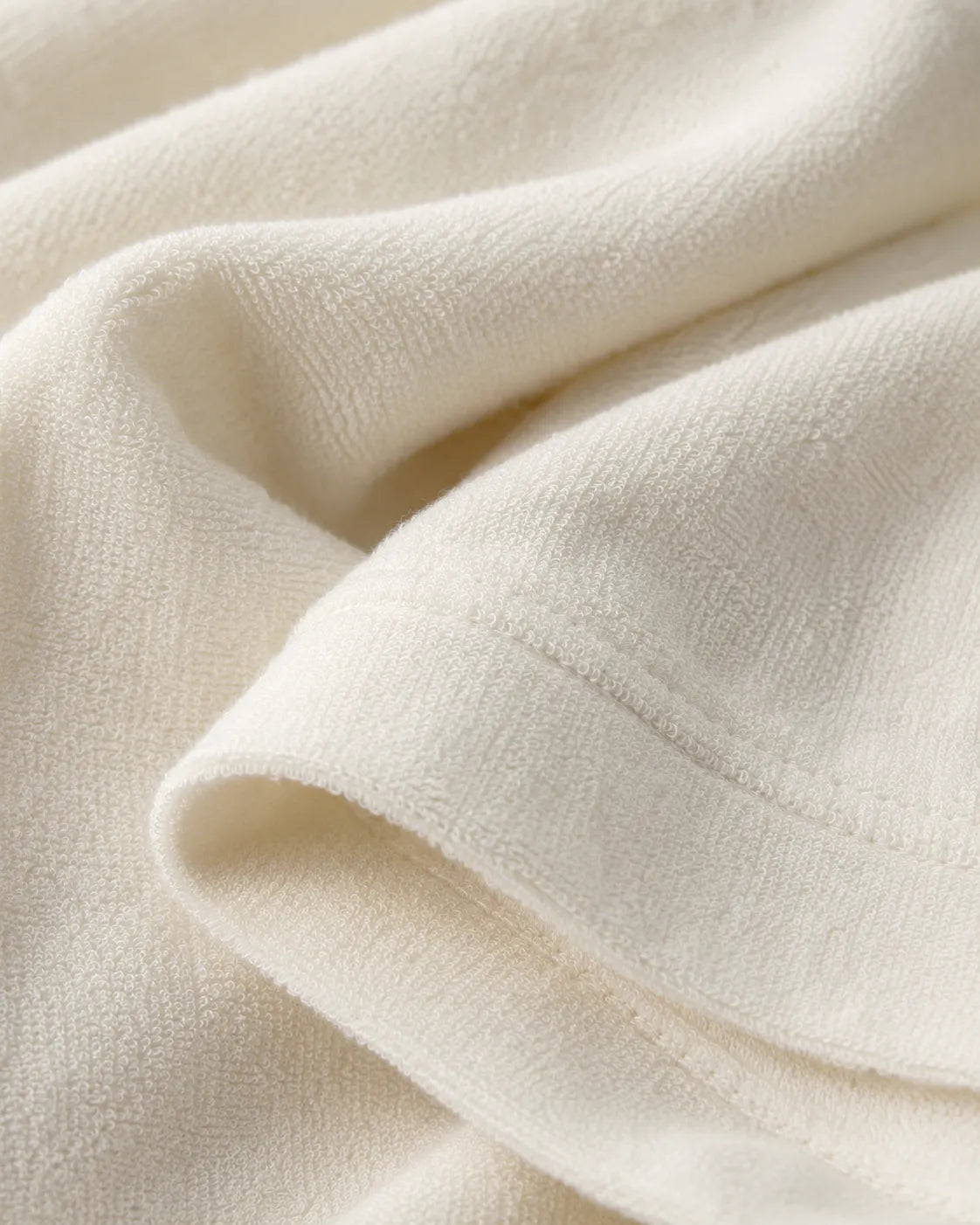 An extreme macro close-up shot focusing on the luxurious texture of a premium off-white Terry knit fabric. The image highlights the soft, looped pile texture, detailed stitching along the hemline, and the heavy drape and natural folds of high-quality streetwear garment material.