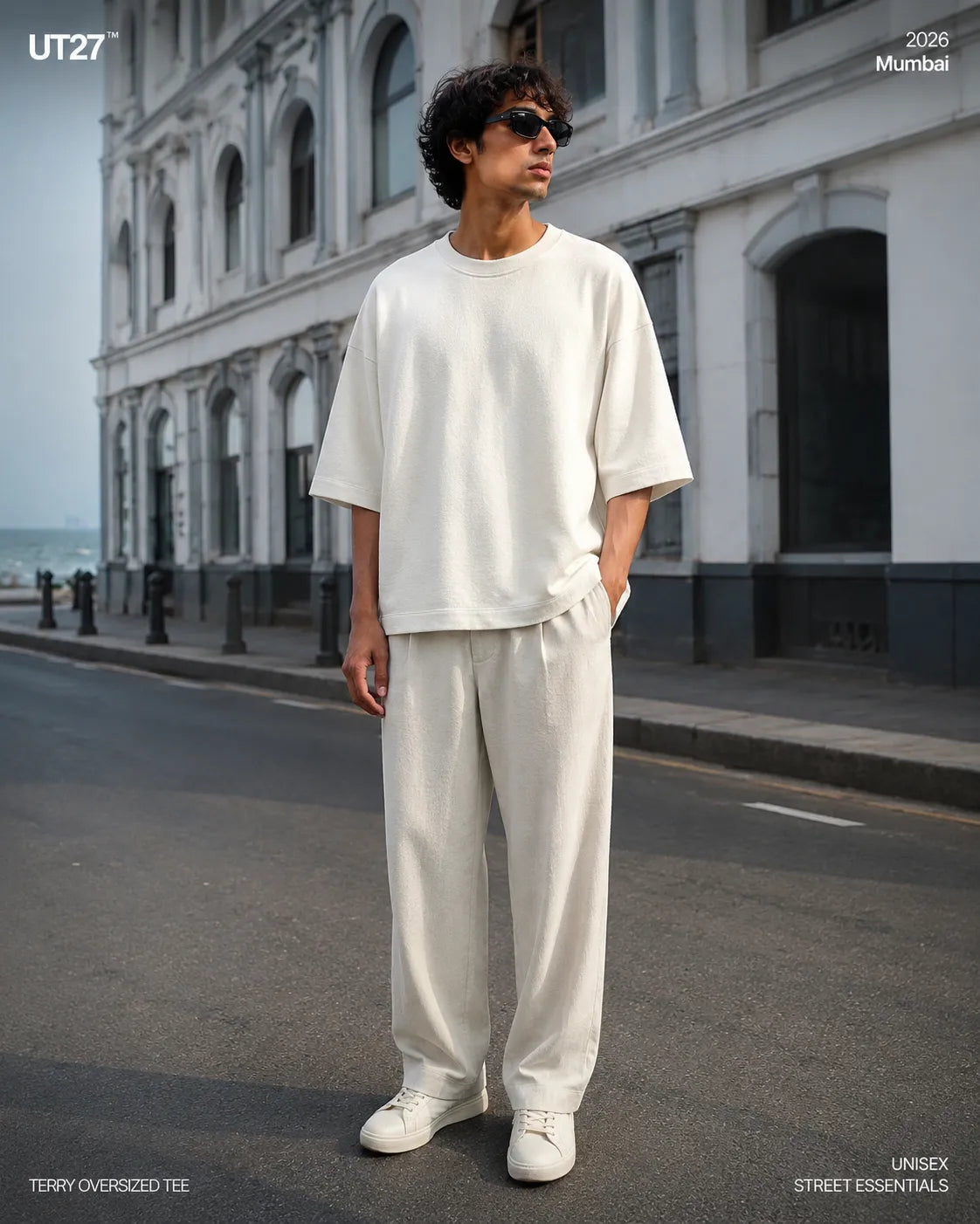 Full-length fashion editorial shot of a male model standing on an urban street in Mumbai, wearing an off-white heavyweight Terry oversized t-shirt and matching relaxed-fit wide-leg trousers. He is wearing dark sunglasses and white sneakers, posing against a classic colonial white building facade. High-end streetwear lookbook photography by UT27.