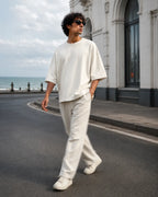 A dynamic street style shot of a model walking along a coastal road next to the sea, captured in motion blur. The model is wearing a premium cream-colored terry knit boxy tee and relaxed trousers set with sunglasses. The background shows a curved street, heritage architecture, and a cloudy sky over the ocean.