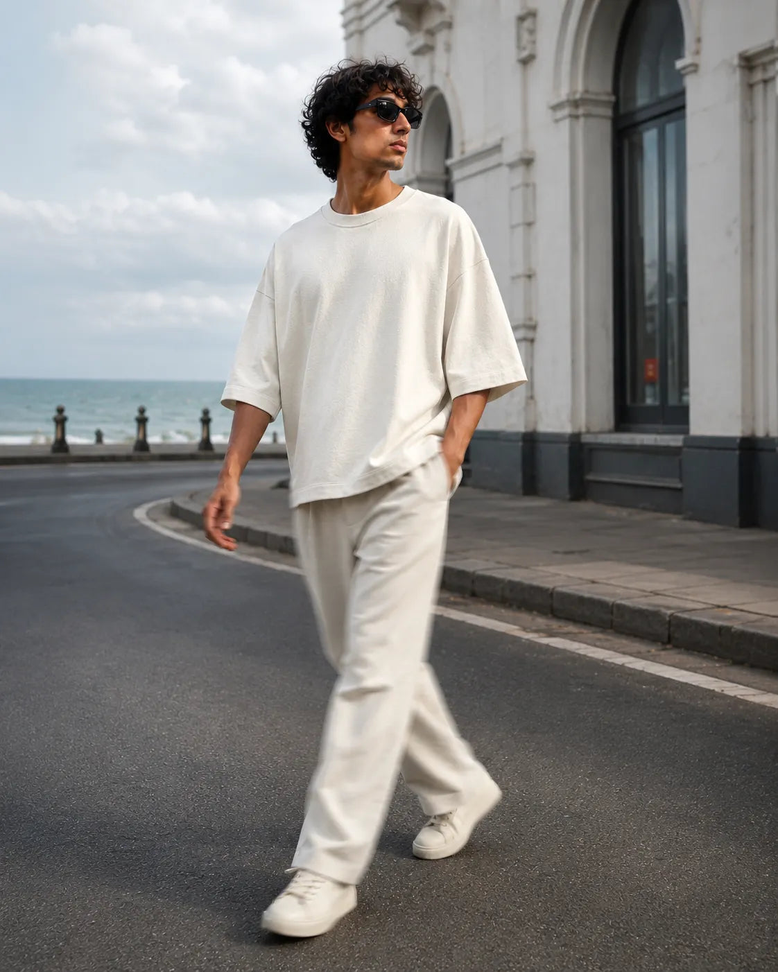 A dynamic street style shot of a model walking along a coastal road next to the sea, captured in motion blur. The model is wearing a premium cream-colored terry knit boxy tee and relaxed trousers set with sunglasses. The background shows a curved street, heritage architecture, and a cloudy sky over the ocean.